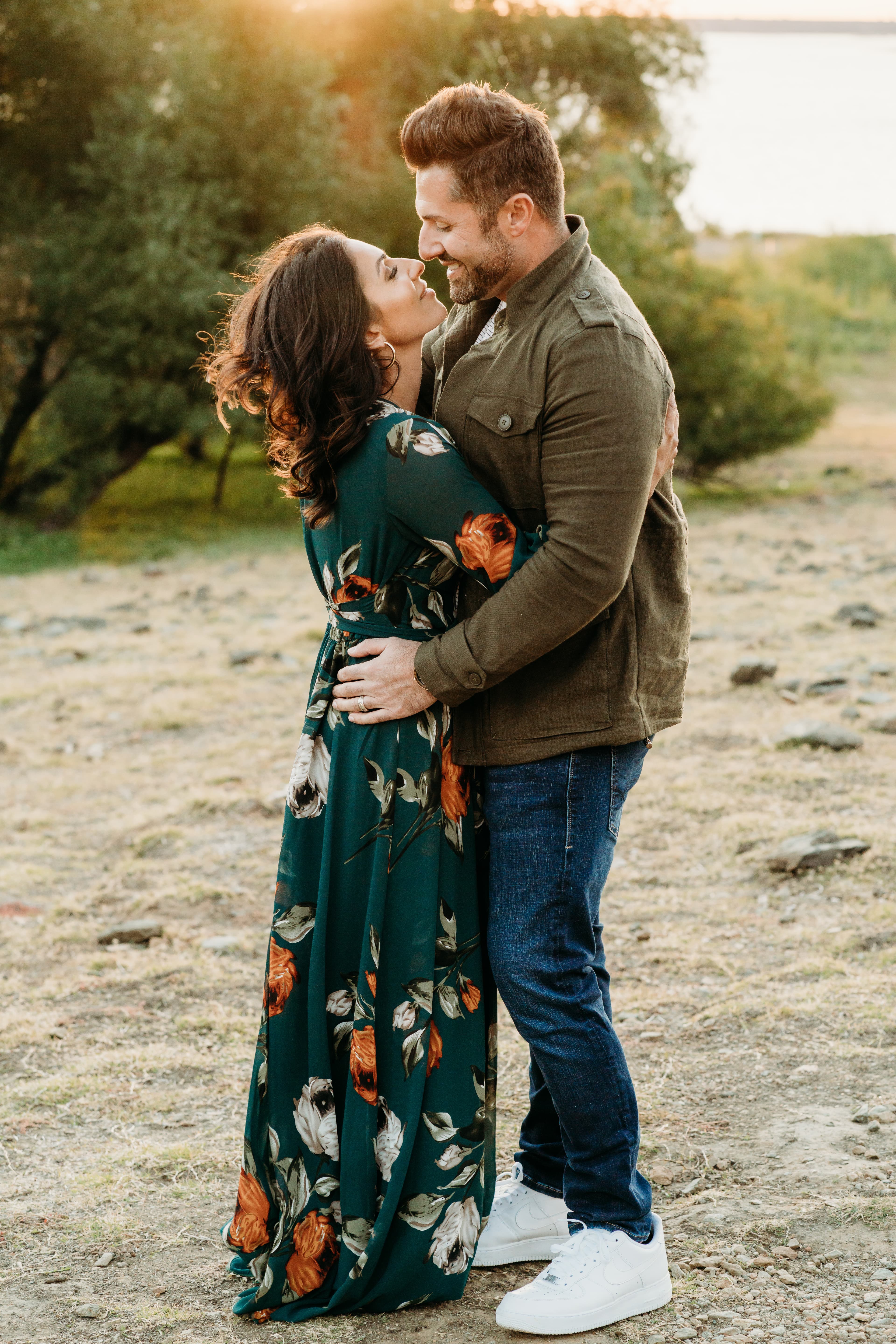 Couple stands nose to nose smiling, she in a green floral dress, golden sunset light
