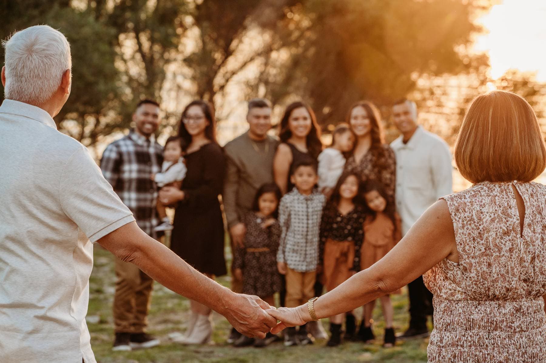 Grandparents seen from behind hold hands as they face their smiling multi-generational family gathered outdoors at sunset