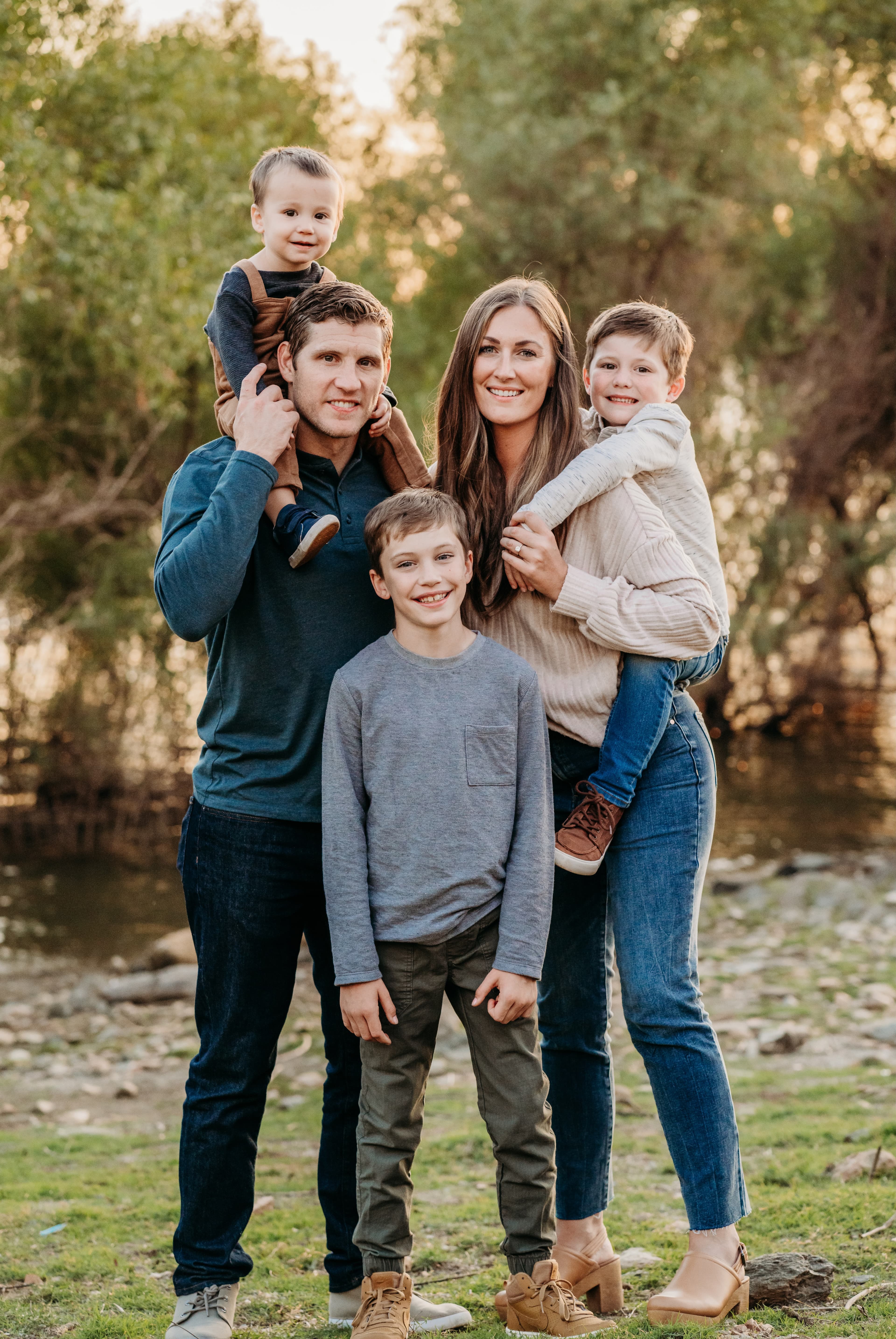 Family of five plays together by a pond at golden hour, children carried on their parents' shoulders and hips
