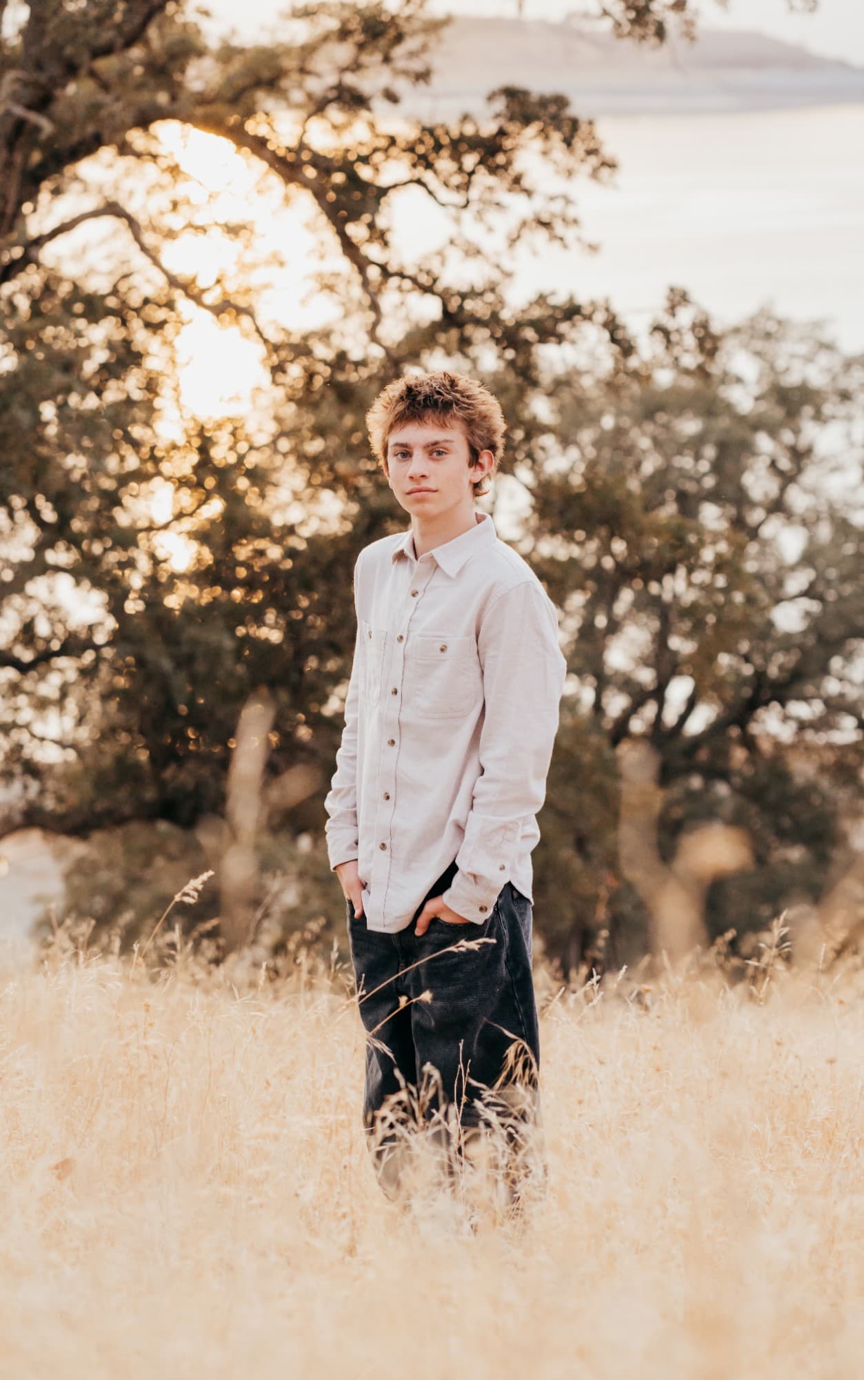 Teen boy in a white button-down stands in a golden field overlooking a lake