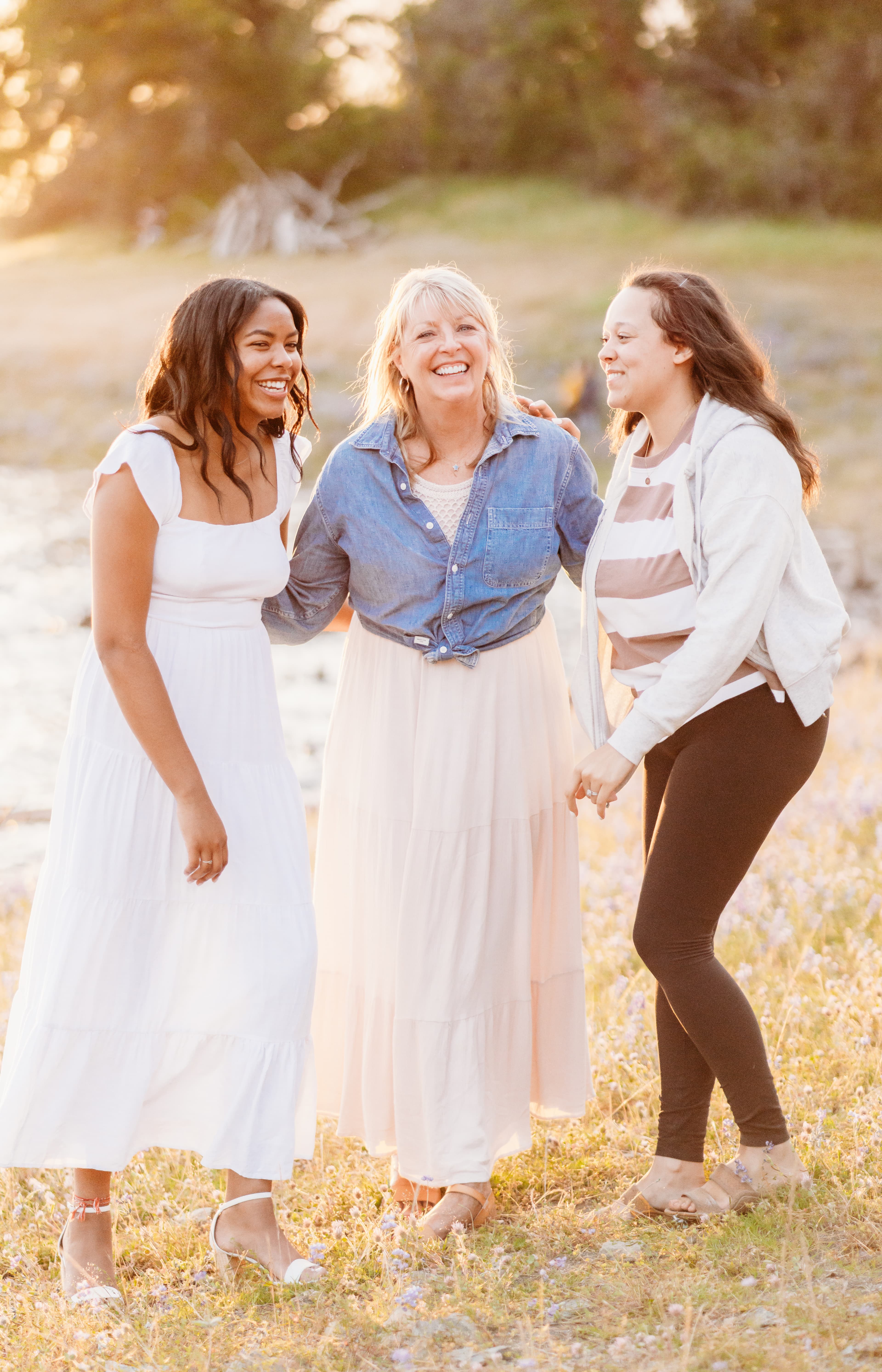 Three women in white, blush, and denim laugh together in a wildflower field