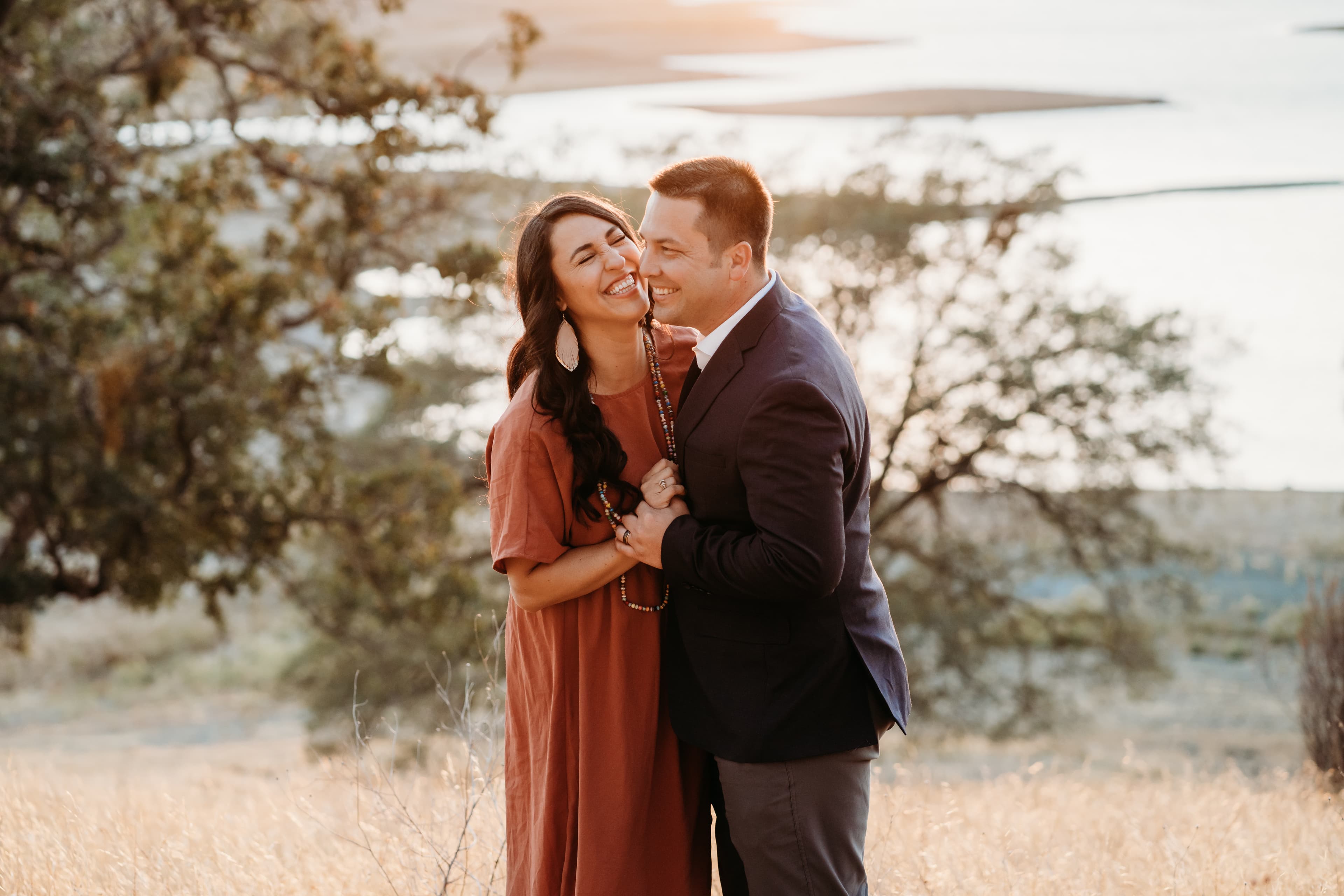 Couple laughs cheek to cheek, she in rust, he in navy, lake at sunset