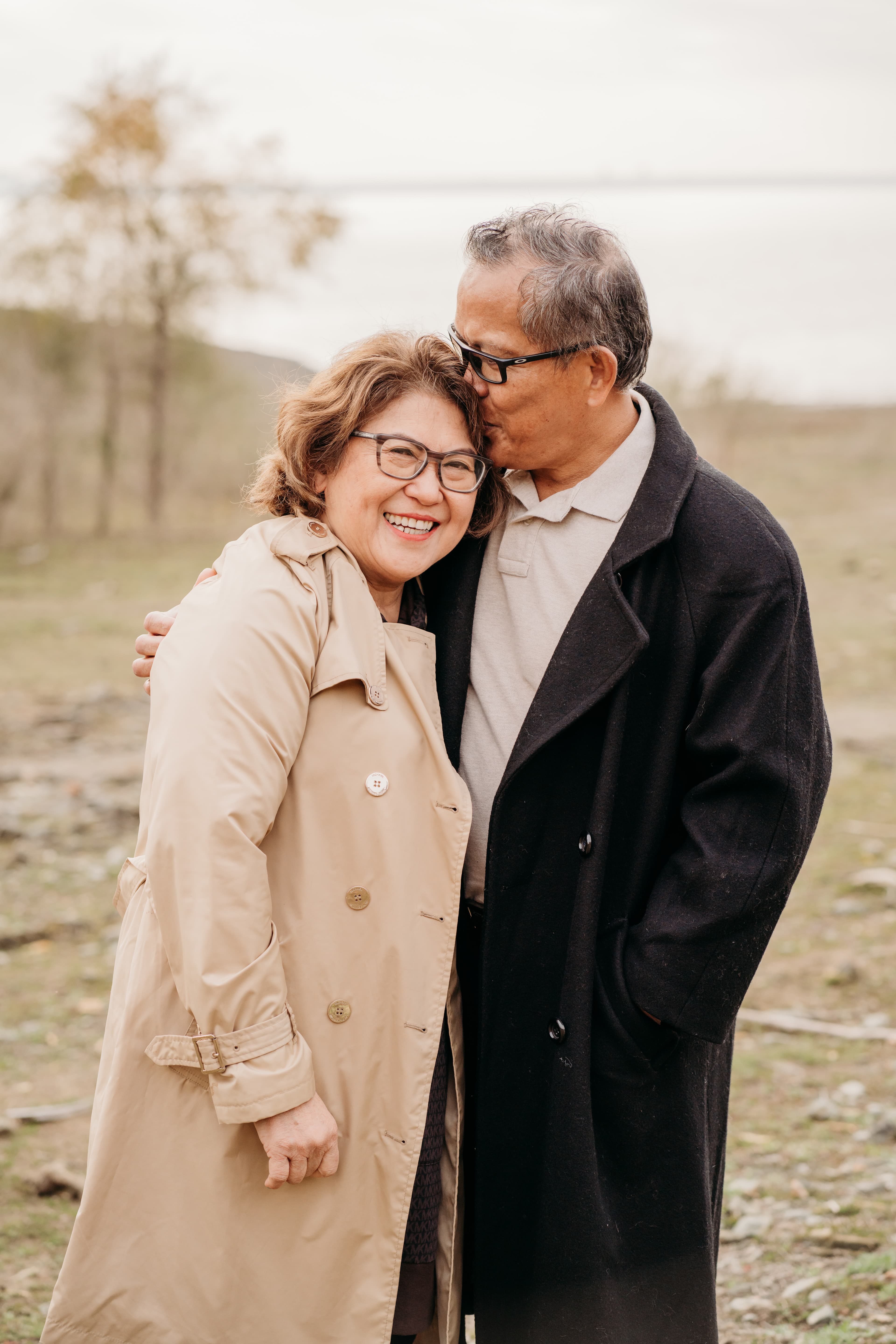 Older man kisses his smiling wife's temple as they embrace outdoors near a lake