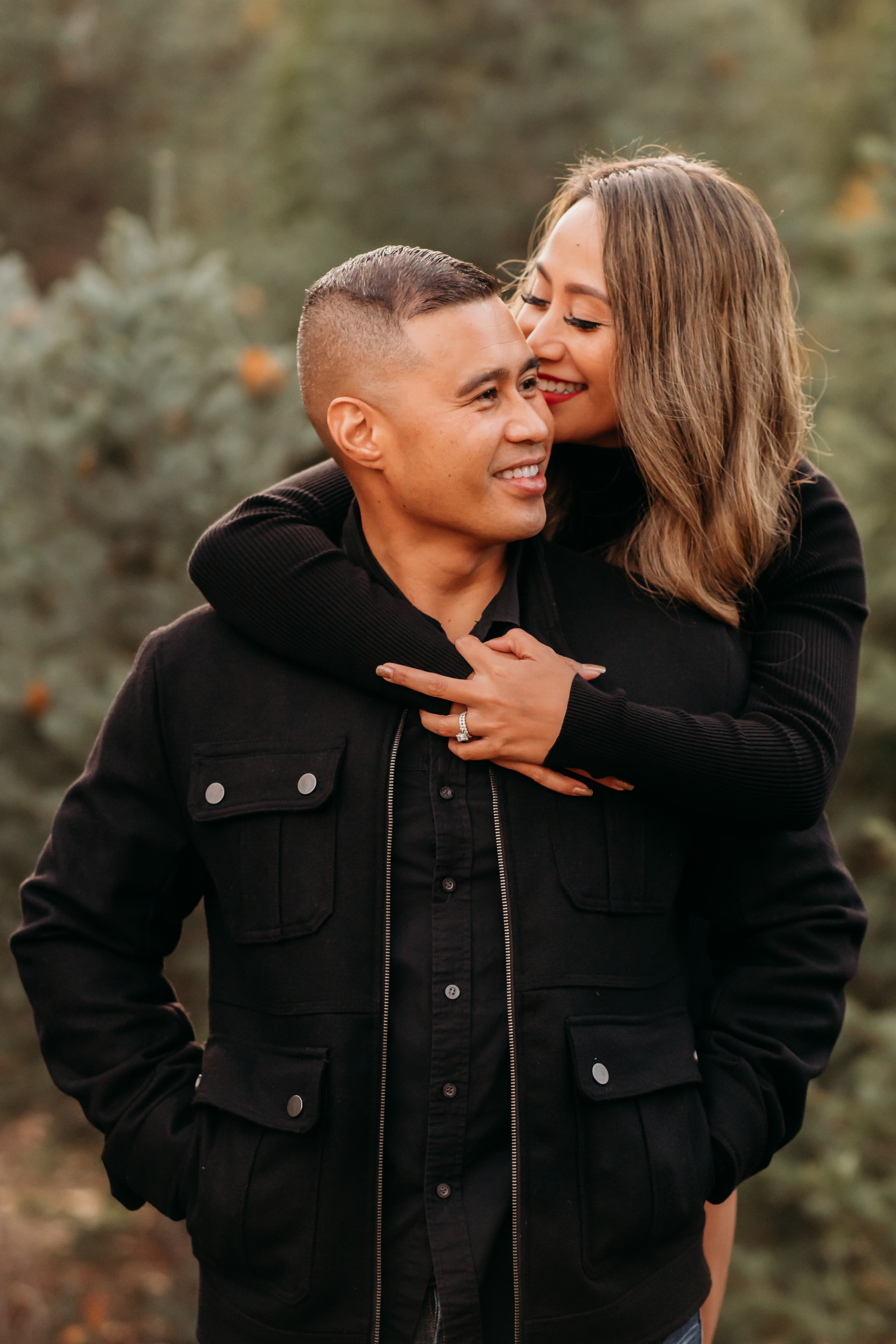Woman hugging her partner from behind, both in black, smiling in a tree farm