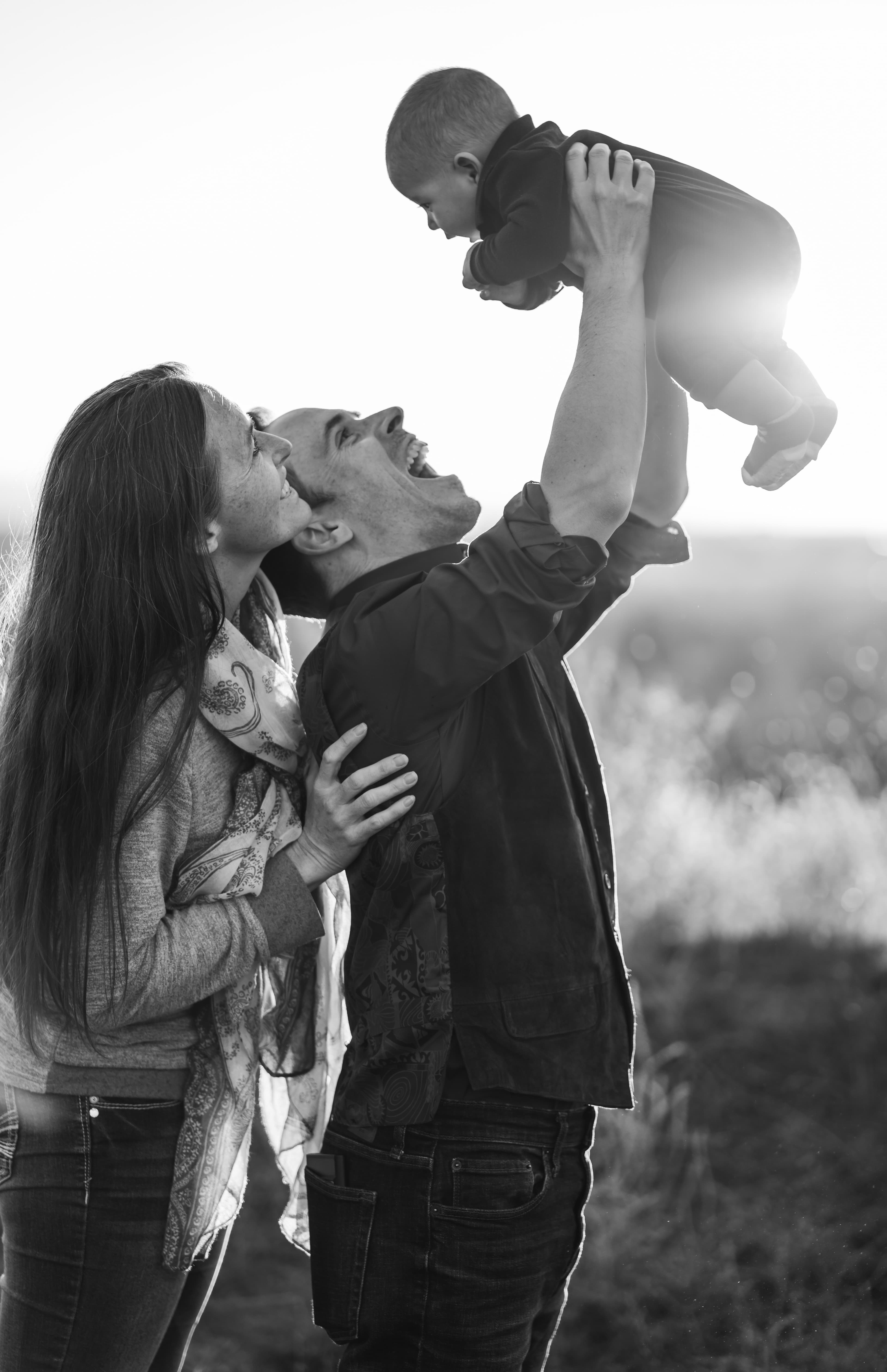 Black-and-white photo of a father laughing as he holds his baby high overhead while the mother smiles on
