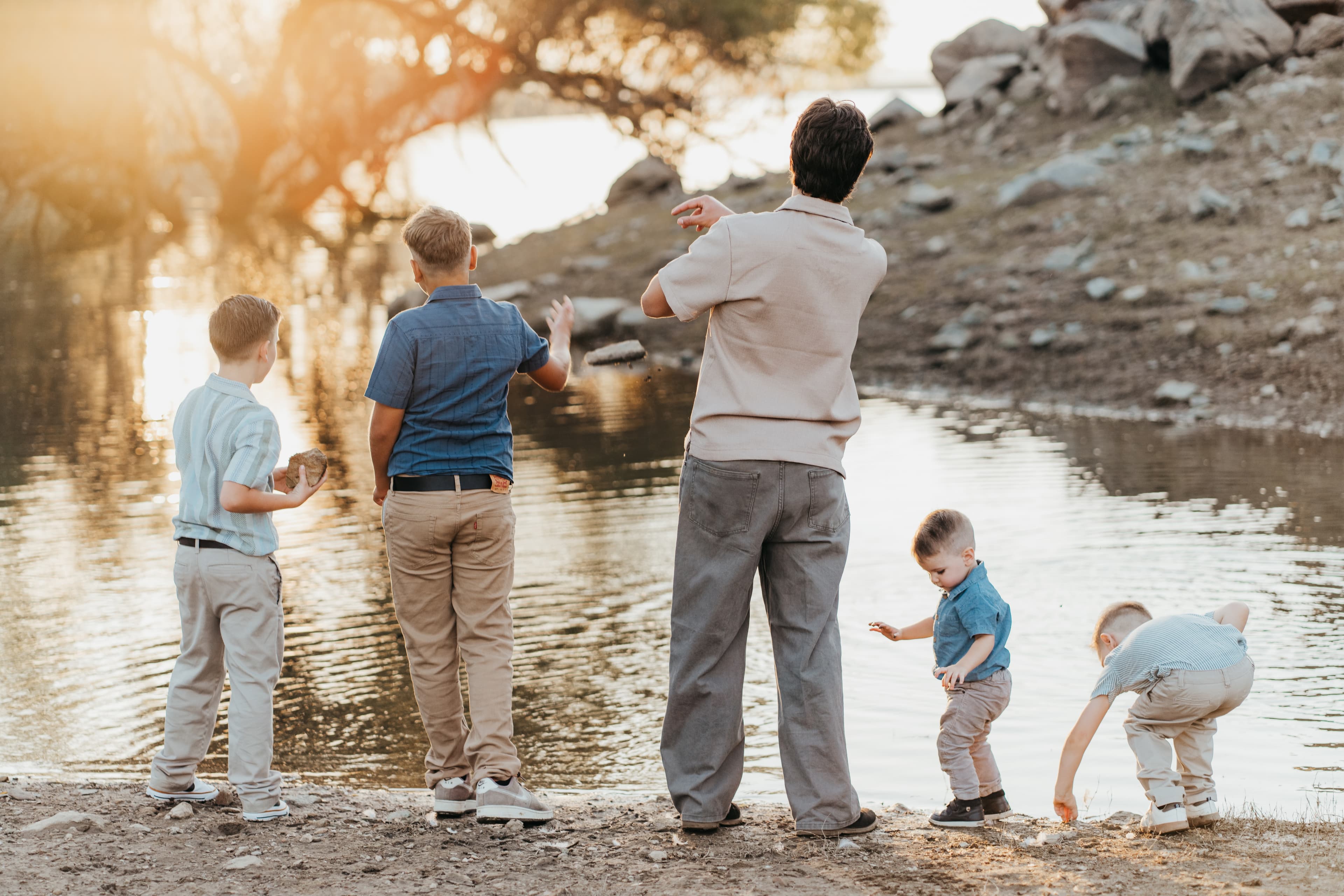 Dad and four sons throw rocks into a lake at sunset, backs to camera