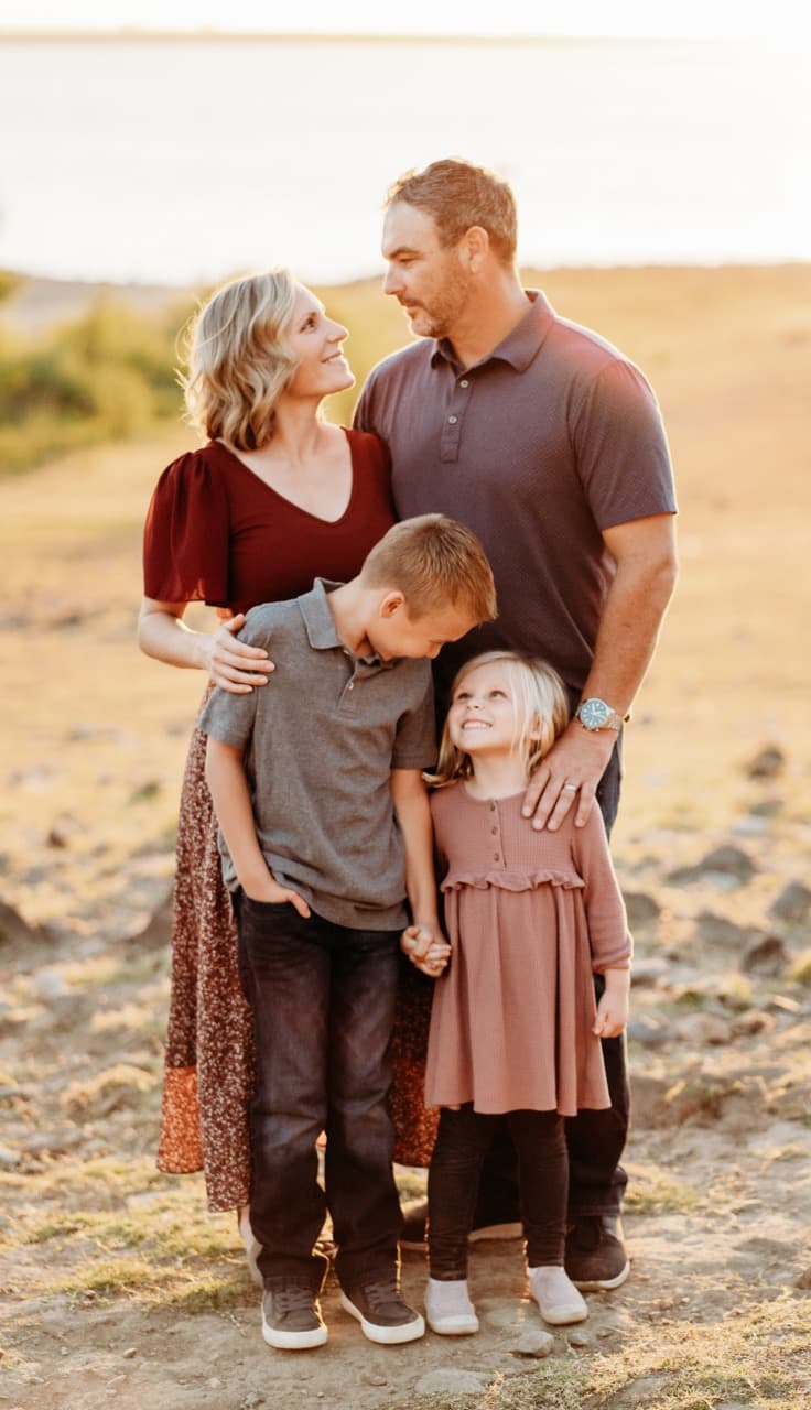 Family of four embraces, parents smiling at each other, kids close by, lake at sunset