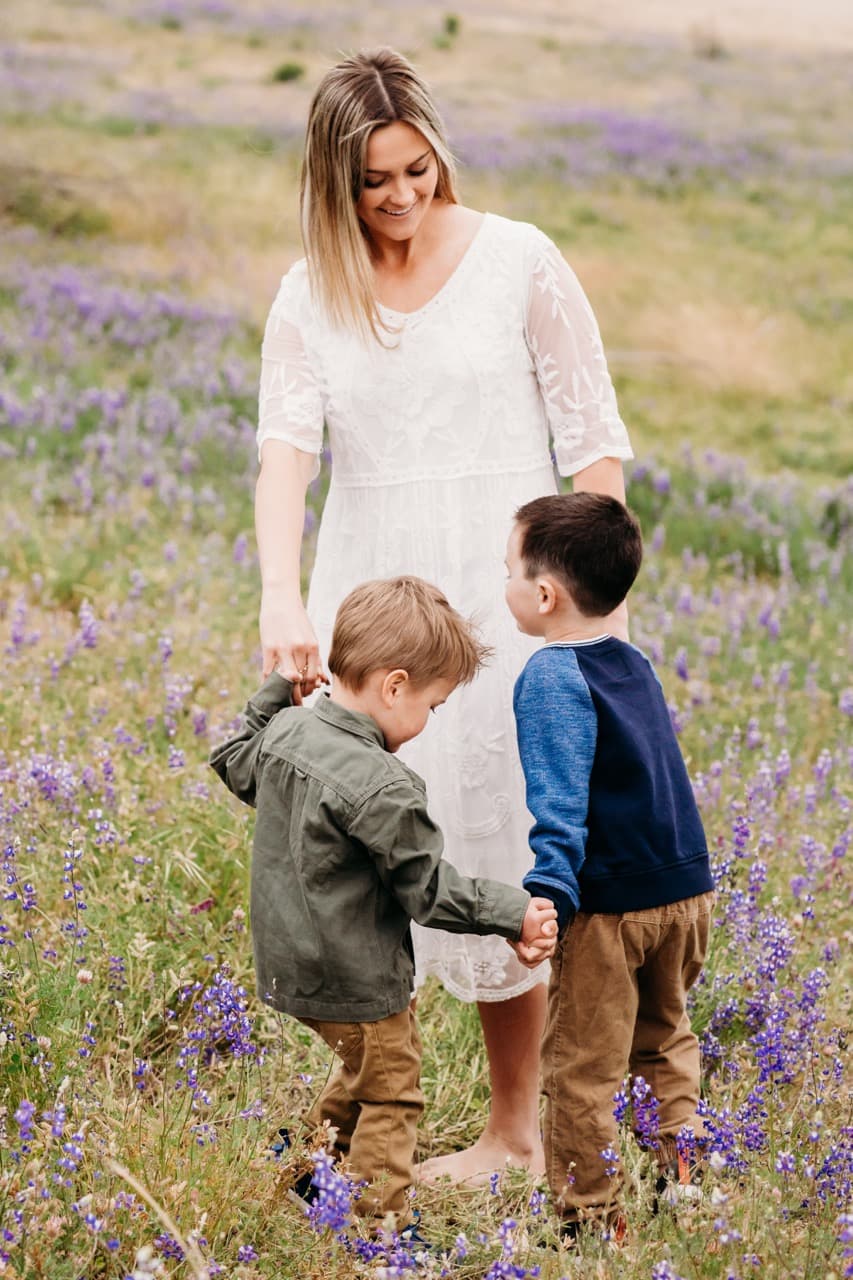 Mother in a white lace dress walks hand in hand with two boys through a lupine field