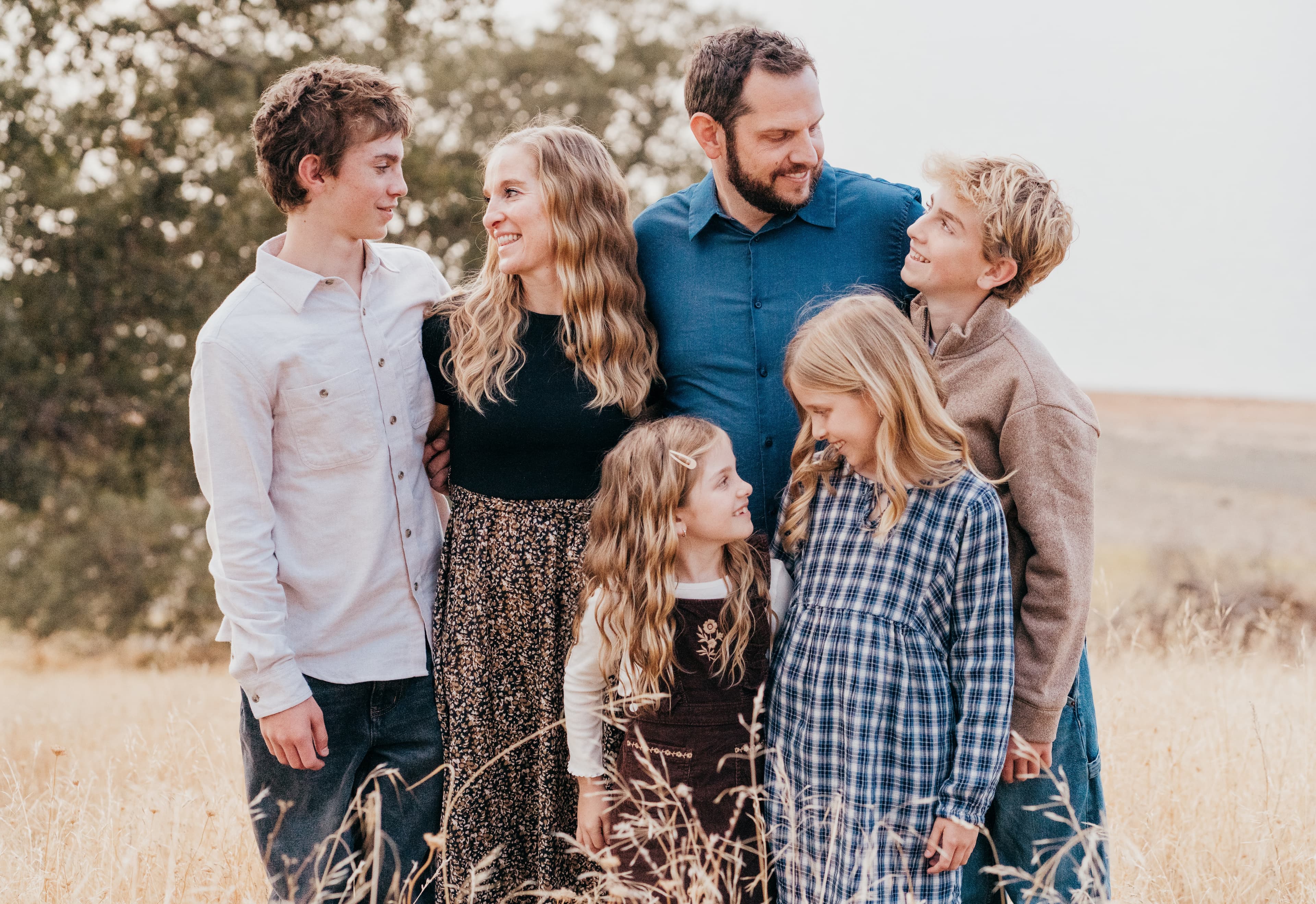 Family of six stands together in a dry golden grass field beneath an oak tree