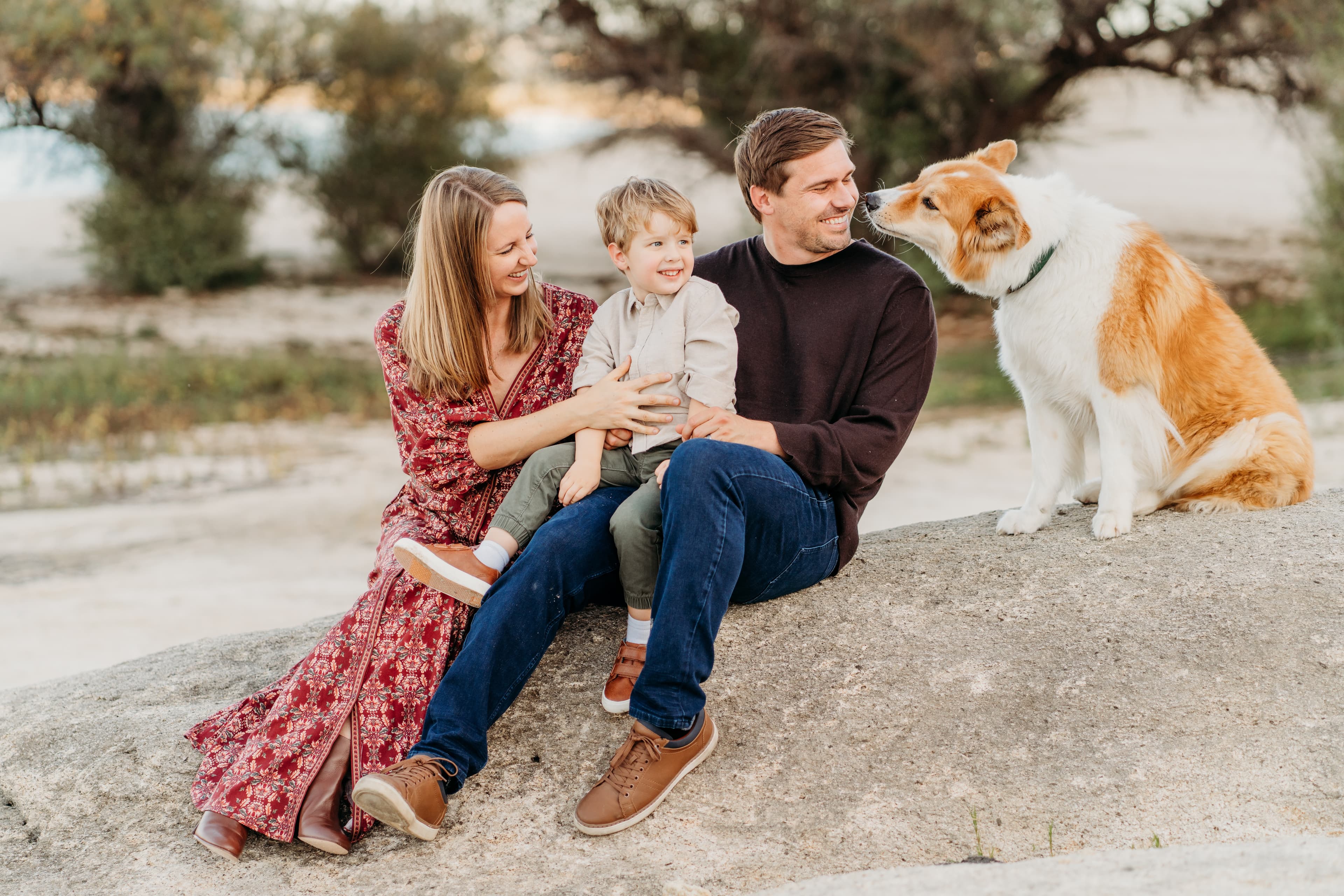 Family of three sits on a granite rock as their dog nuzzles dad's face, lake behind