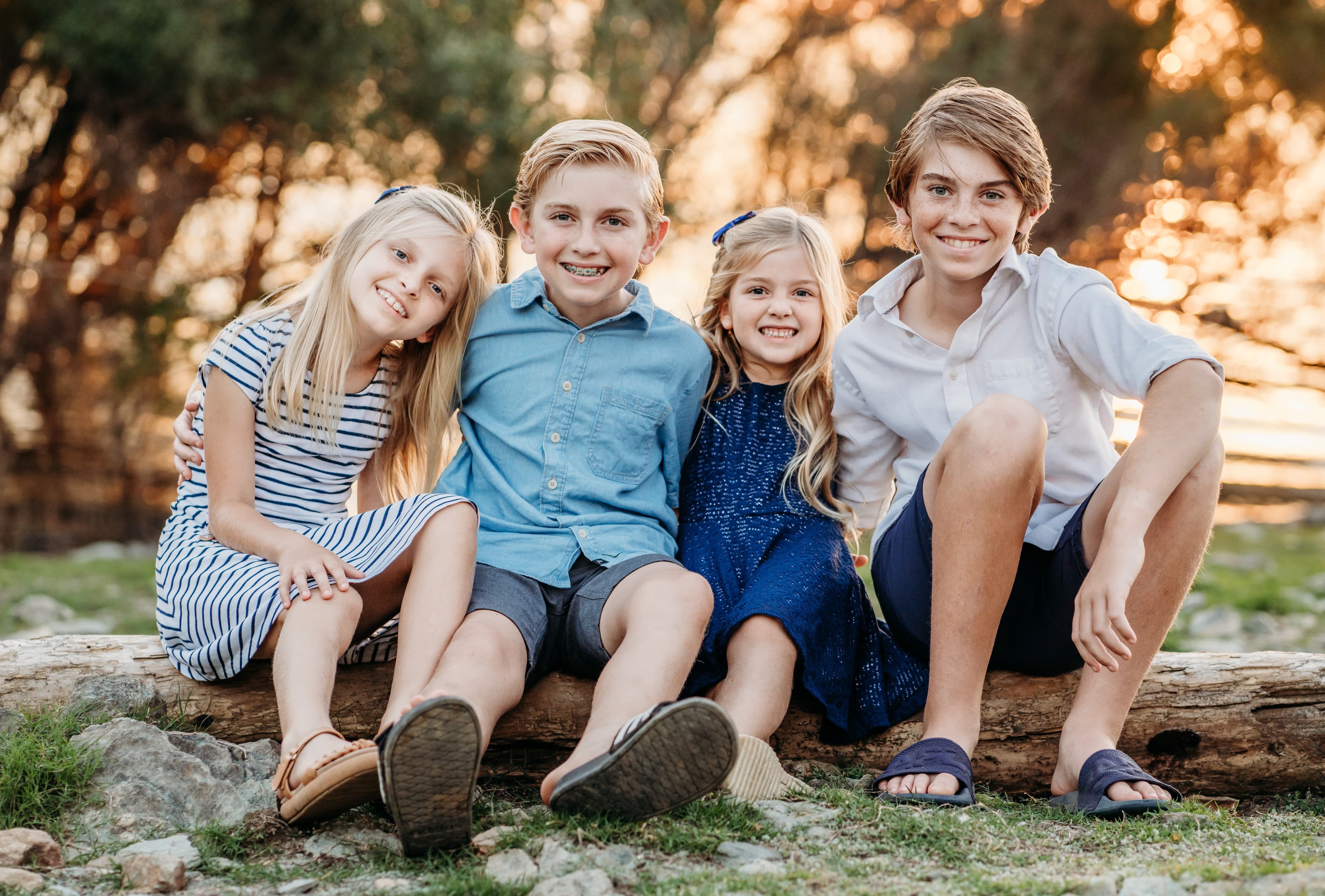 Four smiling siblings sit together on a log in warm golden-hour light