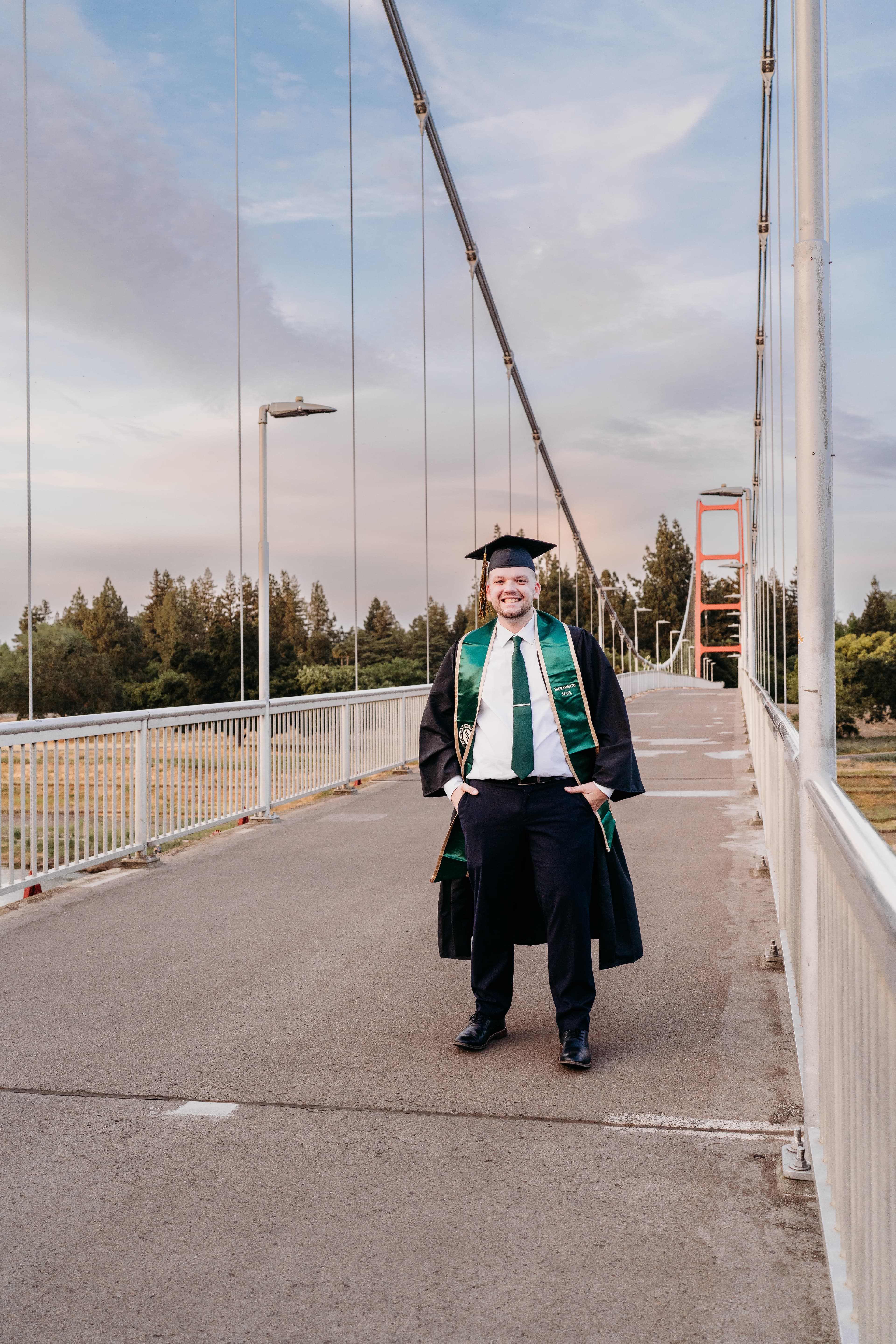 Young man in a cap and gown smiles on a pedestrian bridge at sunset