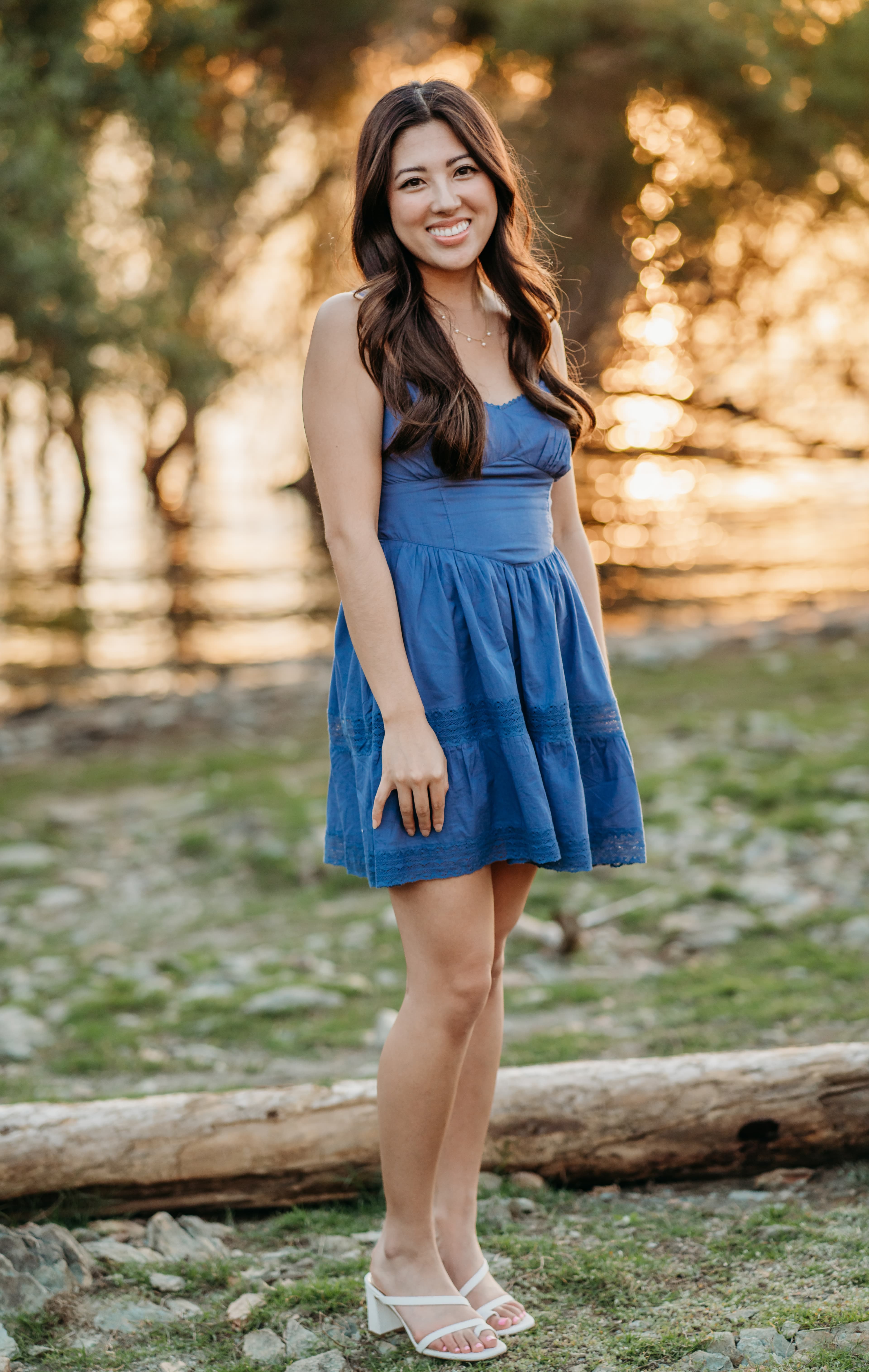 Young woman in a blue dress smiles brightly while standing by a log at the lakeshore