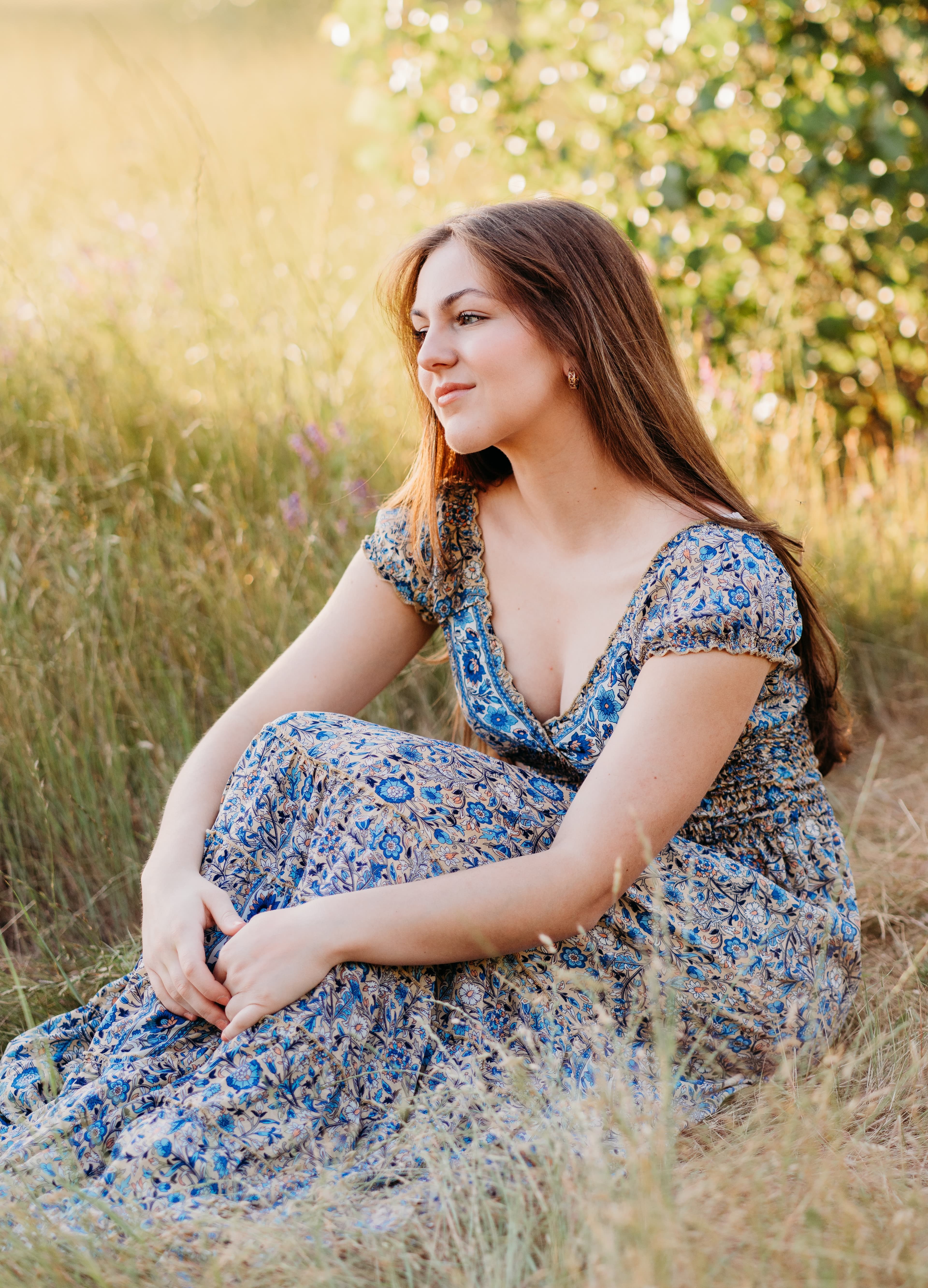 Young woman in a blue floral dress sits in tall golden grass, gazing off to the side