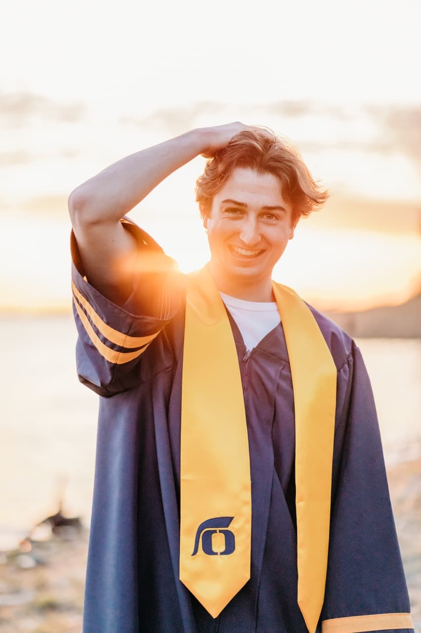 Young man in a navy cap and gown poses alone by a lake at sunset