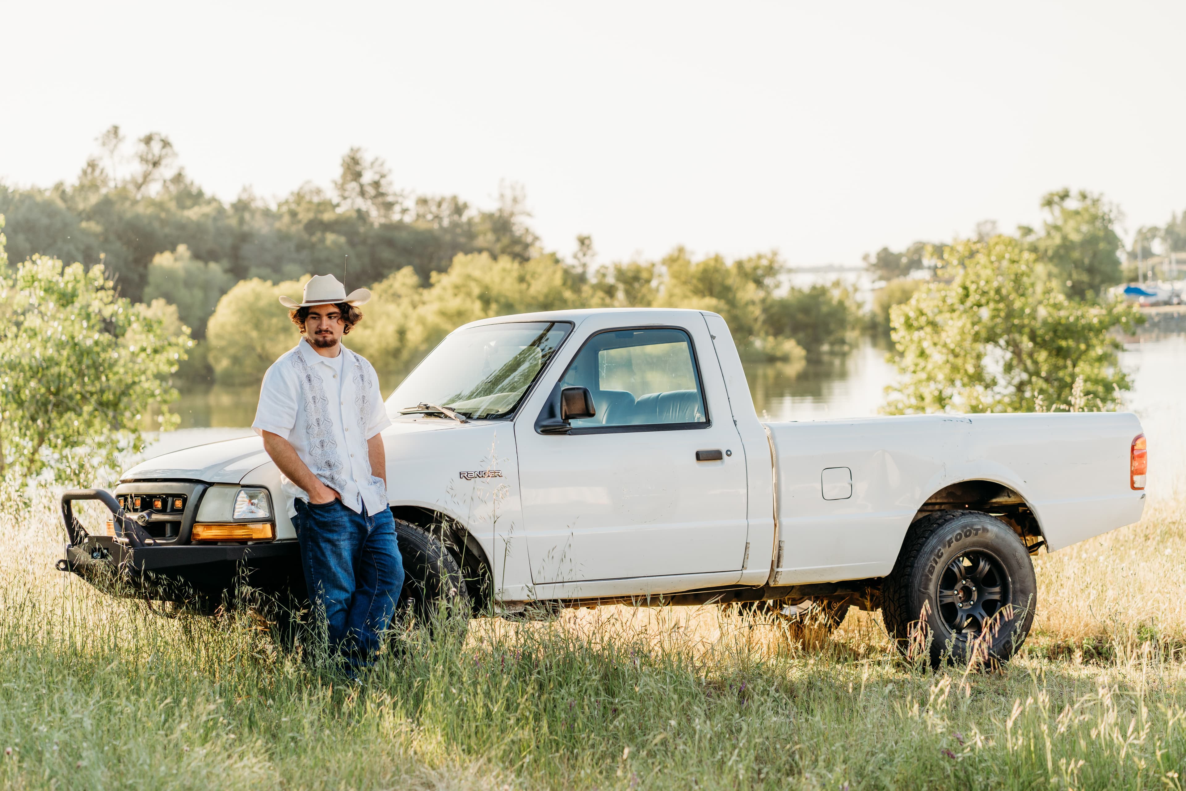 Young man in a cowboy hat leans against a white pickup truck beside a lake