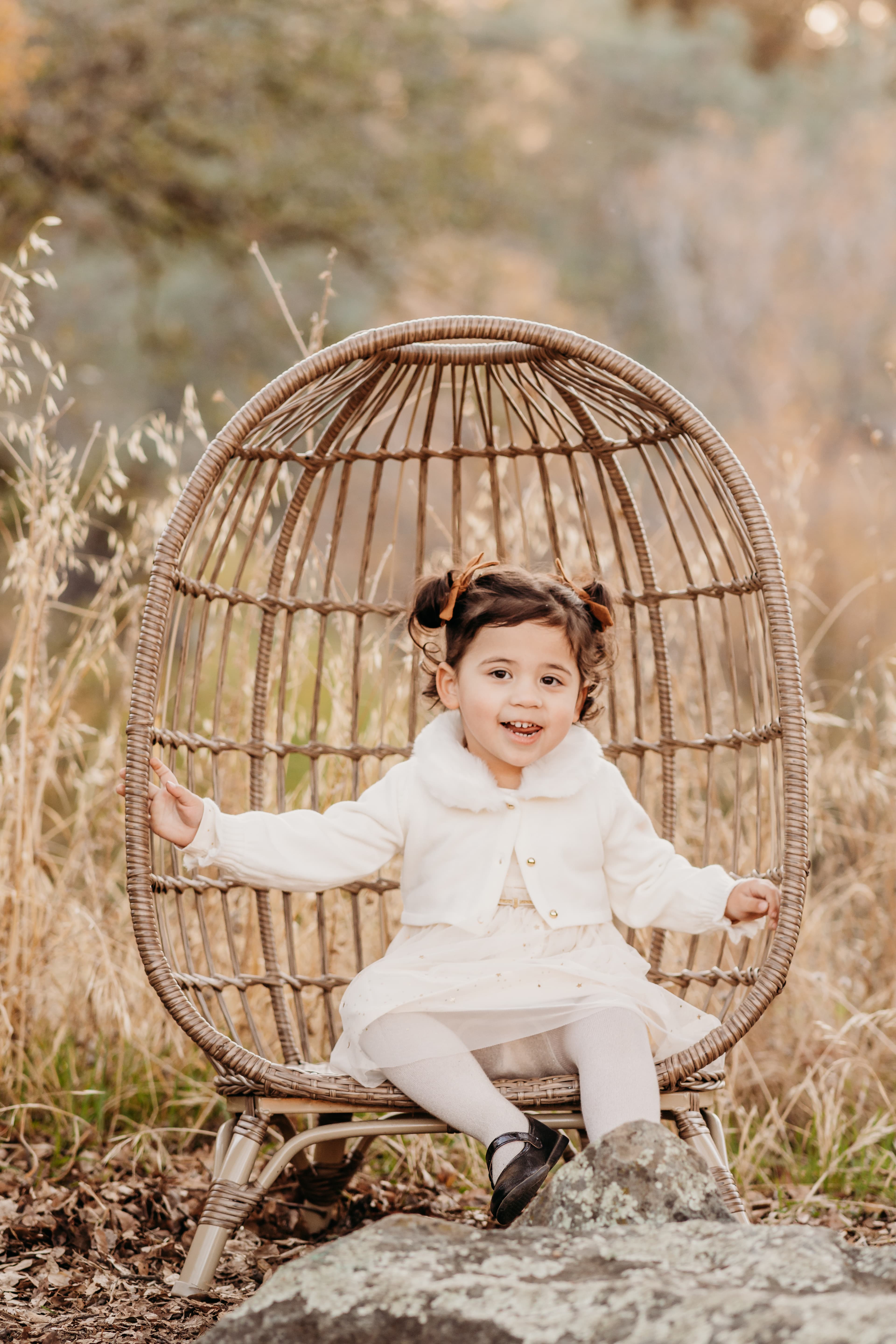 Toddler girl in pigtails sits smiling in a wicker egg chair
