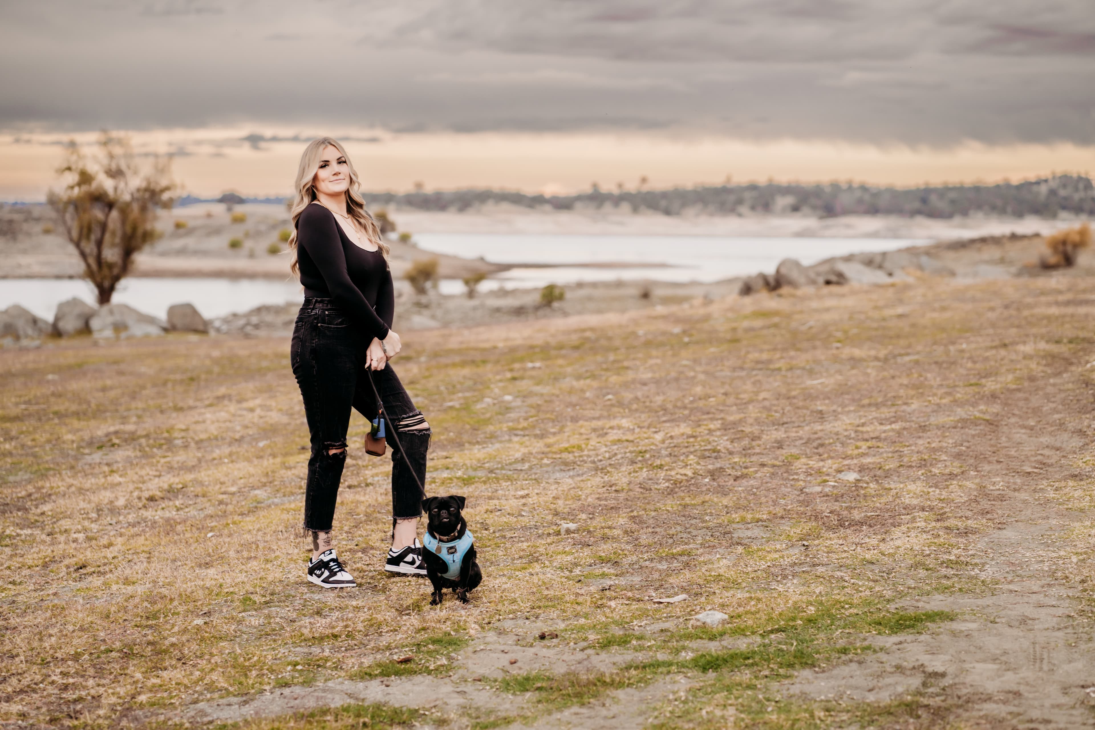 Woman stands on a lakeshore holding the leash of her black pug under a moody sky